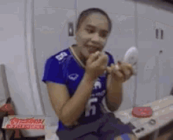 a woman in a blue shirt is sitting on a bed applying makeup .