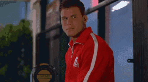 a man in a red jacket is standing in front of a building holding a dumbbell .