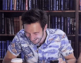 a man wearing a hawaiian shirt is sitting in front of a bookshelf holding a cell phone .