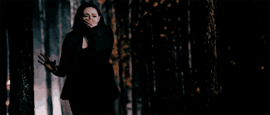a woman in a black jacket is standing in a dark forest covering her mouth