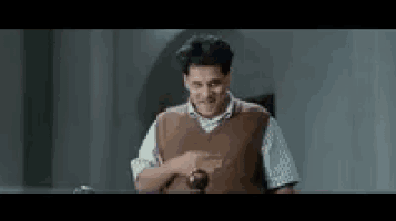 a man in a brown vest is standing in front of a table holding a wooden ball .