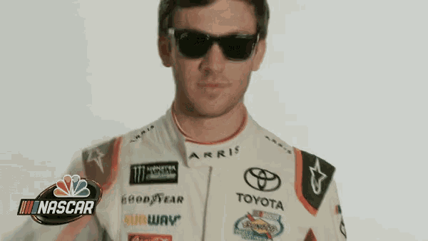 a man wearing sunglasses and a toyota racing suit is standing in front of a nascar logo .