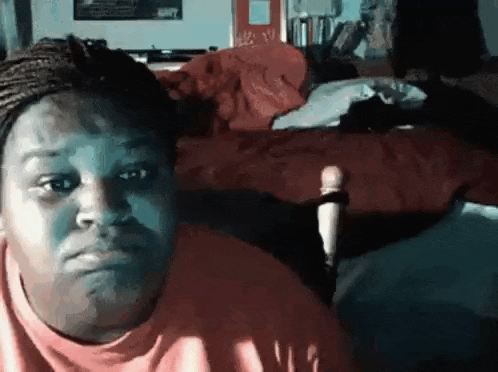 a woman in a red shirt is sitting in front of a bed and making a funny face .