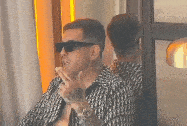 a man wearing sunglasses is smoking a cigarette in front of a mirror .