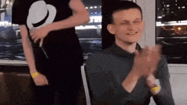 a man in a gray shirt is clapping his hands while another man stands behind him