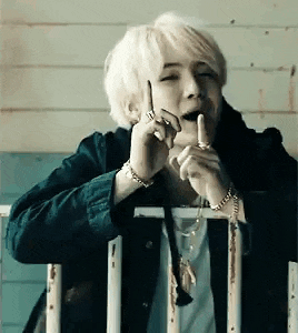 a man with white hair is sitting on a railing talking on a cell phone while holding his finger to his mouth .
