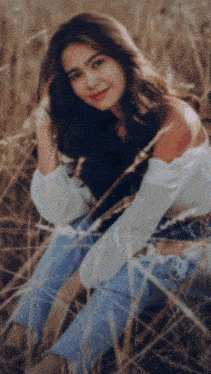a woman in a white off the shoulder top and blue jeans sits in a field of tall grass