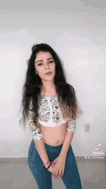 a woman in a crop top and jeans stands in front of a white wall .