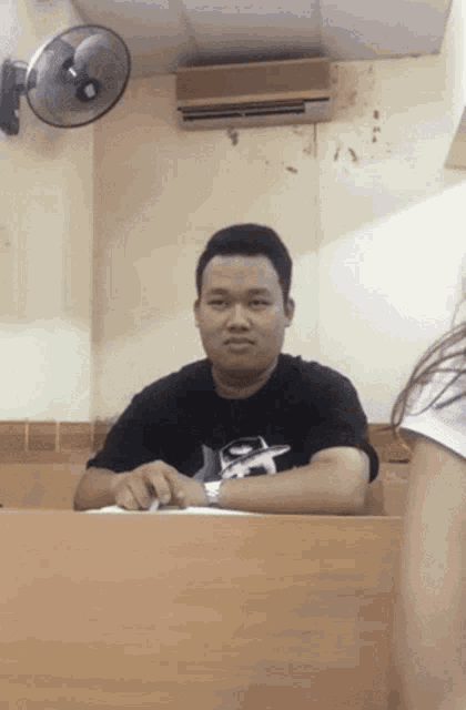 a man wearing a nike shirt sits at a desk with a fan on the wall behind him