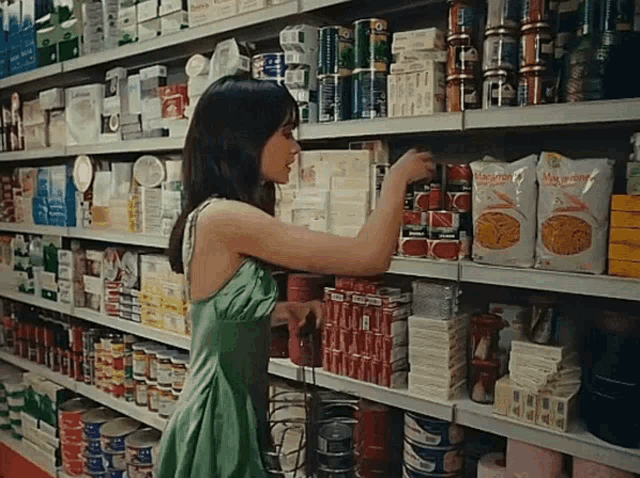 a woman in a green dress is reaching for a bag of macaroni and cheese in a grocery store