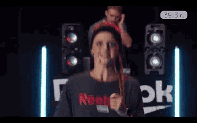 a woman wearing a reebok shirt stands in front of a speaker