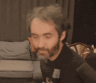 a man with a beard is sitting in front of a couch with a chair in the background