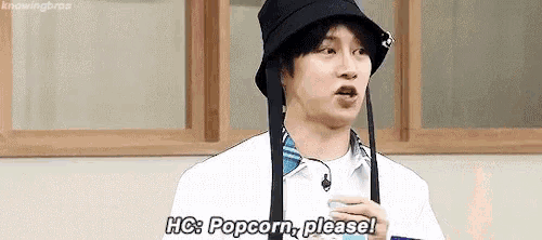 a man wearing a hat is holding a cup of popcorn and saying `` popcorn , please '' .