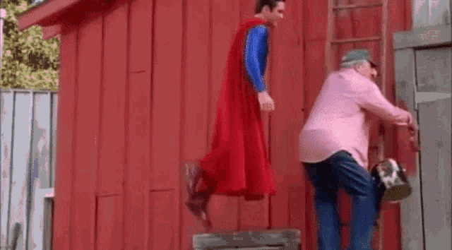 a man in a superman costume is flying in the air next to a man in a pink shirt and a ladder .