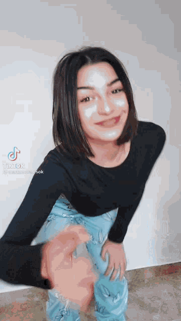 a young woman in a black shirt and blue pants is standing in front of a white wall .