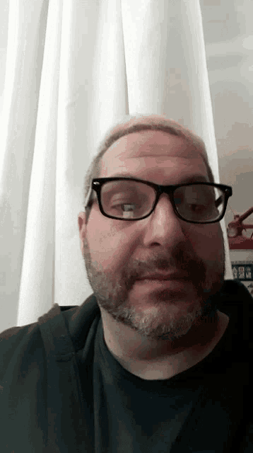 a man with glasses and a beard looks at the camera with a white curtain behind him