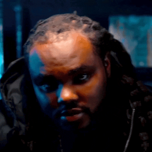 a man with dreadlocks and a beard looks at the camera with a serious look on his face