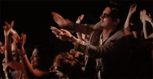 a man in a suit and sunglasses is standing in front of a crowd of people with his arms outstretched .