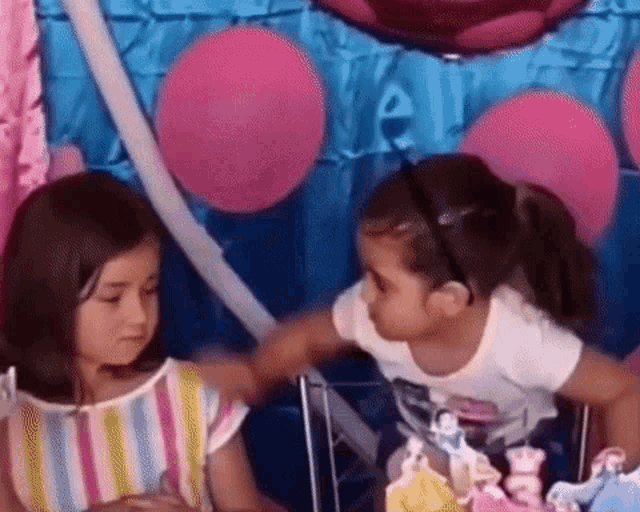 two little girls are standing next to each other at a birthday party and talking to each other .