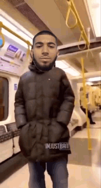 a man with a beard is standing on a train with his hands in his pockets .