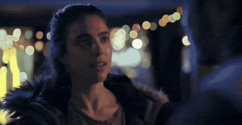 a woman in a fur hooded jacket stands in front of a blurry background of lights