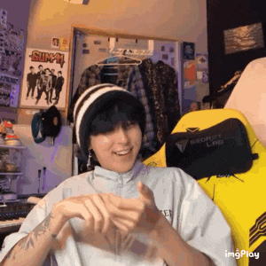 a person wearing a beanie and a white shirt is clapping their hands in a room with a yellow chair .