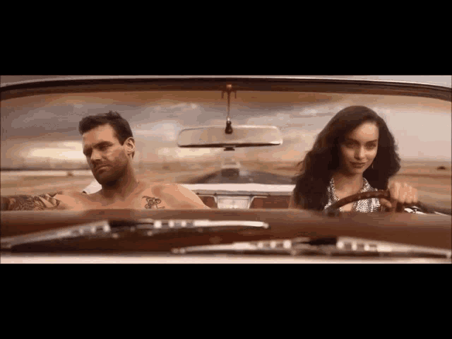 a man with a tattoo on his arm is driving a car with a woman behind him