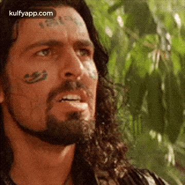 a man with a beard and a tattoo on his face is standing in front of a tree .