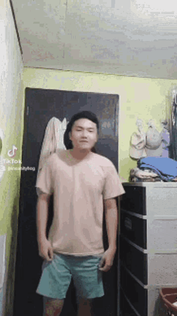 a man in a pink shirt and blue shorts is standing in a room in front of a dresser .
