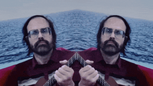 a man with glasses and a beard is playing a guitar in front of the ocean