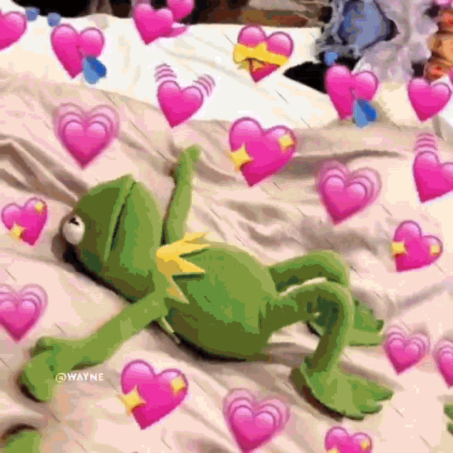 kermit the frog is laying on a bed surrounded by pink hearts and emojis .