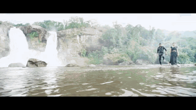 a man and a woman are walking across a river near a waterfall