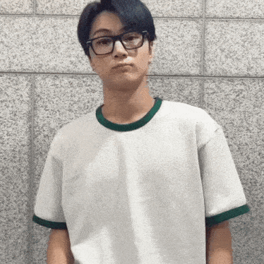 a young man wearing glasses and a white t-shirt is standing in front of a brick wall .