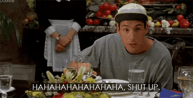 a man in a hat is sitting at a table with glasses of water and flowers and says shut up