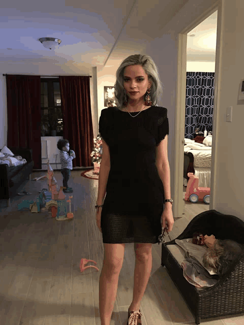 a woman in a black dress stands in a living room with a baby in the background