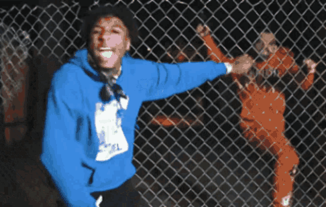 a man in a blue hoodie stands in front of a chain link fence with his tongue out