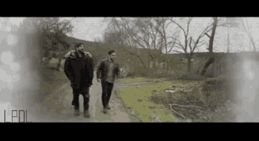 two men are walking down a dirt road in a forest .
