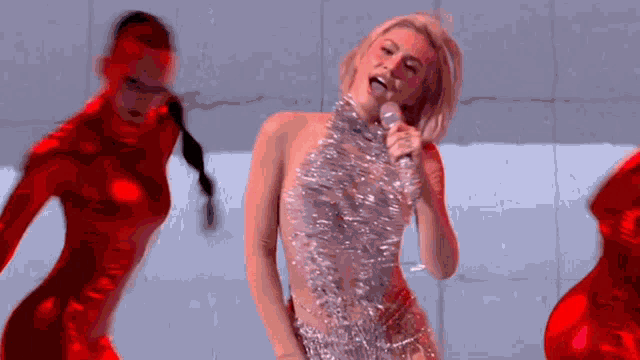 a woman in a silver dress is singing into a microphone while two women in red costumes are dancing behind her .