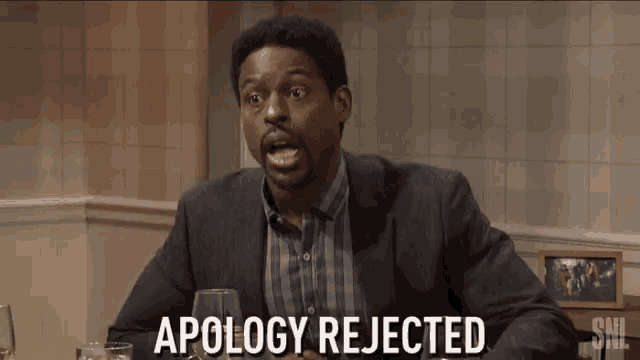 a man in a suit and plaid shirt is sitting at a table with a glass of wine and says apology rejected .