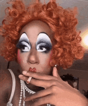 a drag queen with red hair is smoking a cigarette and wearing a pearl necklace .
