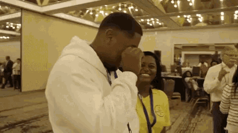a man in a white hoodie is covering his face with his hand while a woman in a yellow shirt is smiling .