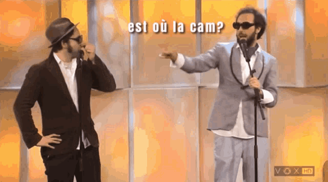 two men standing in front of a microphone with the words est où la cam written above them