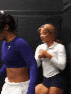 two women are standing next to each other in a room . one of the women is wearing a blue crop top .