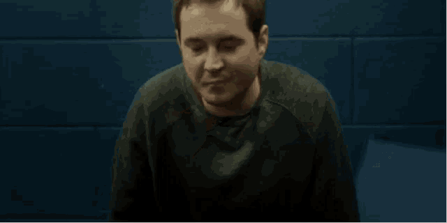 a man in a black sweater is sitting in front of a blue wall in a dark room .