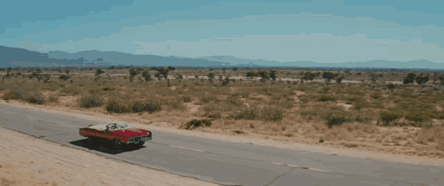 a red car is driving down a desert road with mountains in the background