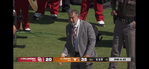 a man in a suit and tie stands on a field with a scoreboard that says oklahoma 20 texas 35