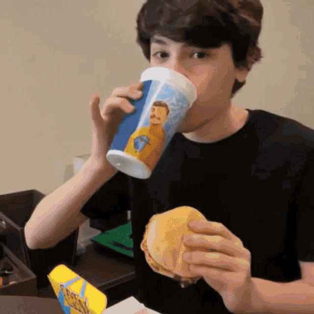 a boy drinking from a cup with a picture of a man on it while eating a hamburger