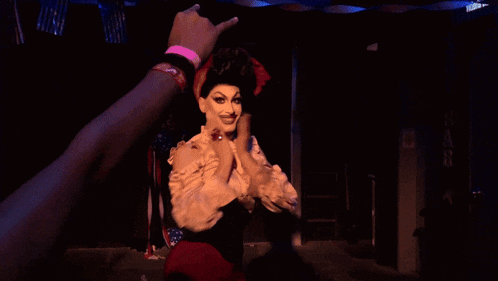 a drag queen stands in a dark room with flags hanging from the ceiling