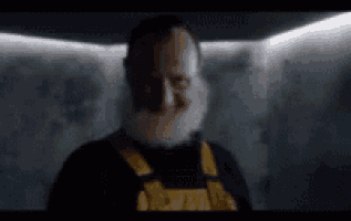 a man with a beard and overalls is standing in a dark room and smiling .
