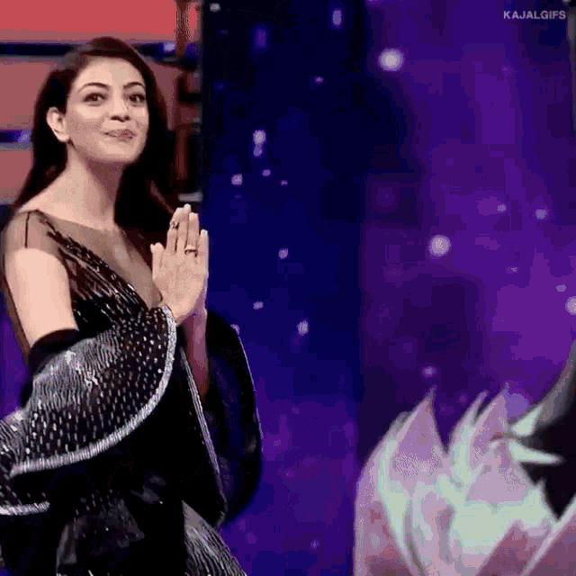 a woman in a black dress is standing in front of a purple background with her hands folded .